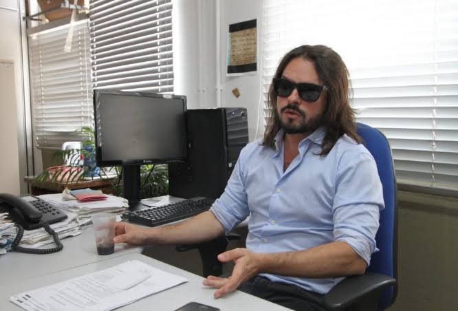 Rafael Giguer está no seu local de trabalho, sentado e com as mãos apoiadas na mesa, ele está de óculos escuros e tem cabelo pelo ombro