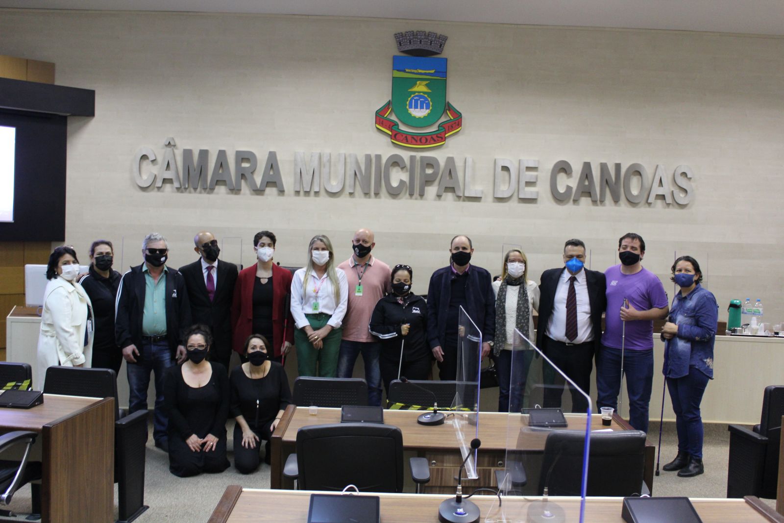 Foto com 15 pessoas, sendo 9 mulheres e 5 homens, eles estão de pé na frente da mesa do plenário da Câmara Municipal de Canoas. Na foto estão representantes da ADEVIC, ONCB e do Executivo Municipal.