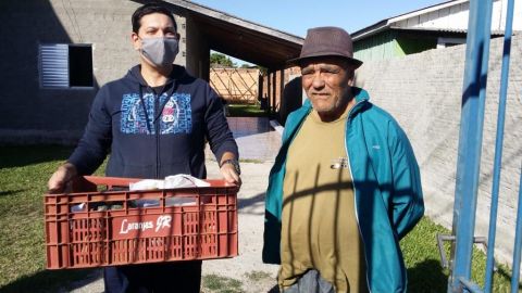 São dois homens em frente a uma residência. O primeiro, é o voluntário da ADEVIC, usa máscara e segura com as duas mãos a caixa de mantimentos.