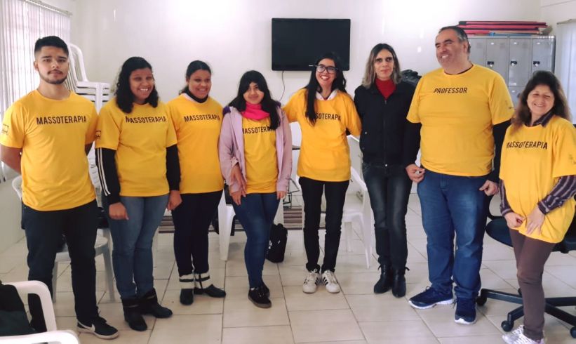 Alunos e o professor da turma de Massoterapia Inclusiva posam para foto. Eles estão em pé, vestidos com as camisetas do projeto na cor amarela.