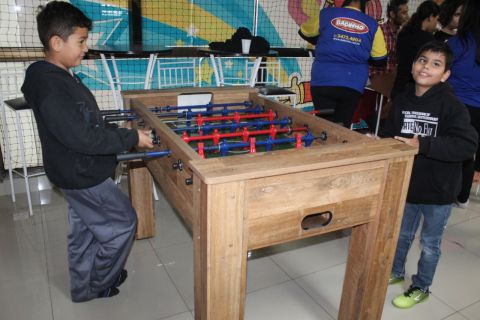 Dois garotos associados da ADEVIC brincam em uma mesa de Fla-Flu. Ambos usam moletom preto, estão em pé, concentrados no jogo