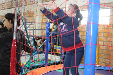 Associada da ADEVIC brinca na cama elástica. A cama é azul, é cercada por uma rede de proteção na cor vermelha. Ao lado da cama, ao fundo, tem uma piscina de bolinhas coloridas. 