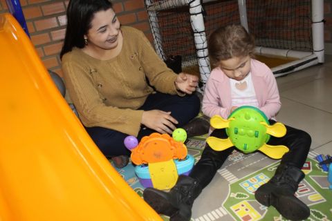 Menina associada da ADEVIC se diverte com brinquedos coloridos. Ela está sentada no chão e ao lado dela uma mulher observa e sorri. 