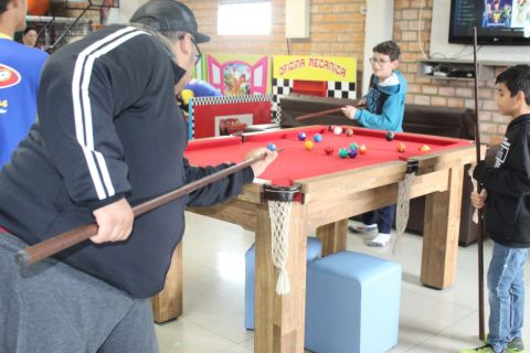 Três garotos estão em pé ao redor de uma mesa de sinuca vermelha, jogando.