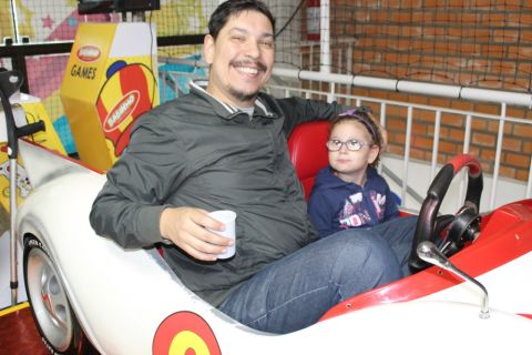 A pequena Eloisa, atendida pela ADEVIC, acompanhada pelo pai, se diverte dentro de um minicarro branco, com assentos vermelhos. O pai olha para a câmera no momento da foto muito sorridente.
