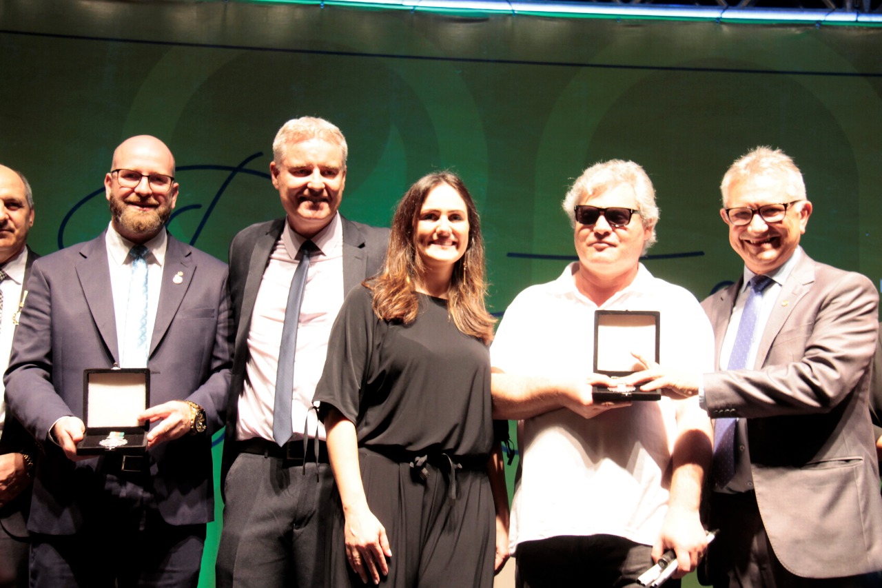 No palco do evento, o prefeito entrega a medalha ao presidente da Adevic, José Guimarães. Ao lado dele estão também a vice-prefeita e diversas outras pessoas.