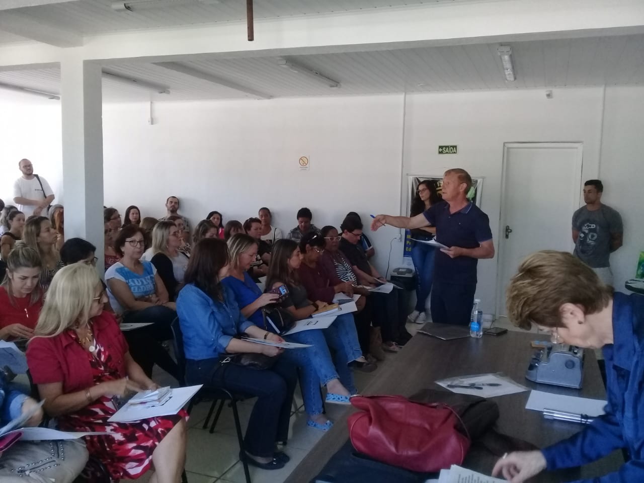 Um grande grupo de pessoas sentadas formam uma plateia no local de realização do workshop. Elas seguram folhas de papel nas mãos e observam o instrutor do workshop, que está em pé diante delas, gesticulando durante sua explicação. 
