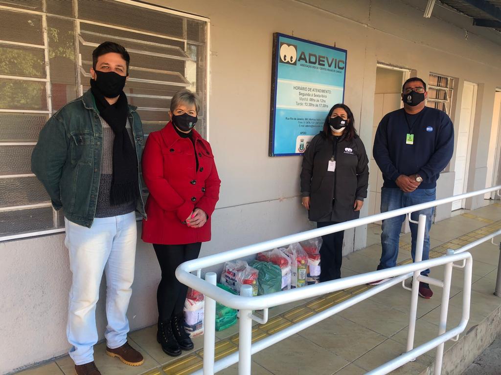 Foto com quatro pessoas de pé, elas estão no corredor de acesso para as demais salas da ADEVIC. Da esquerda para a direita tem um homem (Felipe), de máscara preta, manta preta e veste jaqueta jeans, ao seu lado uma mulher de casaco vermelho (Fernanda), cabelo curto e usando uma máscara preta, no meio estão algumas sacolas de cestas básicas, após tem uma mulher de cabelo pelo ombro (Cíntia), com máscara preta da ADEVIC e com um casaco da ADEVIC e ao seu lado um homem de blusão azul marinho (Alexsandro) e máscara preta.